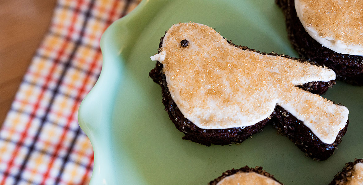 A bird-shaped chocolate cake on a plate rests on a table topped with a patterned tablecloth.