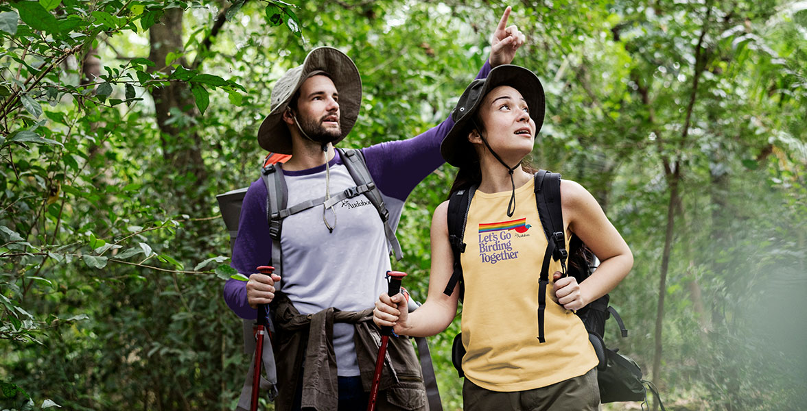 Two people bird in a woodsy area holding hiking poles. The person on the left points to a faraway spot. The person to their right looks to where they're pointing.